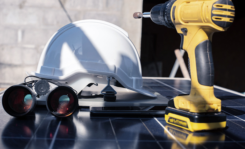 Close-up of a white safety helmet, yellow cordless drill, binoculars, and a tablet placed on a solar panel during installation work.