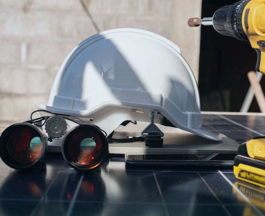 Construction tools including helmet, binoculars, drill and tablet placed on a solar panel at a photovoltaic installation site.