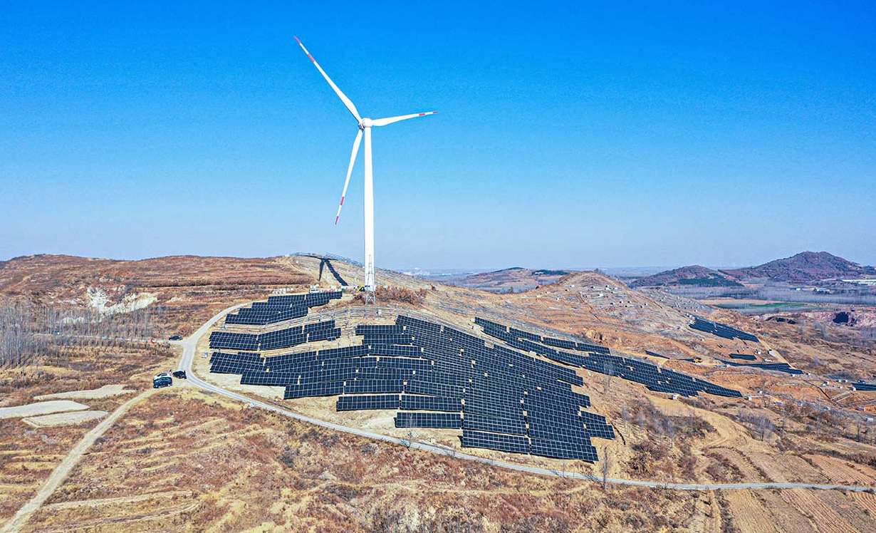 A hybrid renewable energy site featuring both wind turbines and solar panels on a mountainous landscape under a clear blue sky
