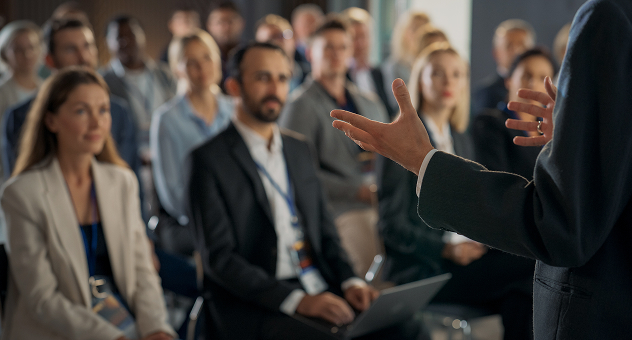 Business speaker addressing an audience at a professional conference, with attendees listening attentively