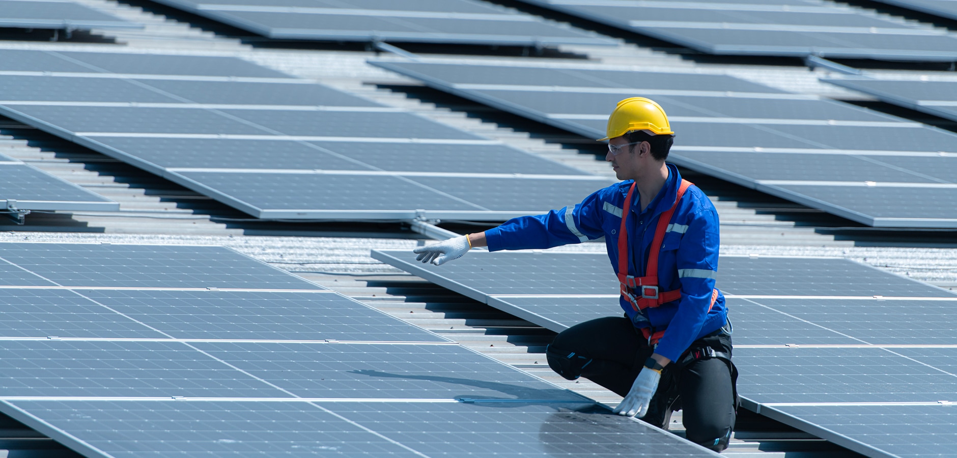 Worker inspecting and maintaining solar panels on a rooftop installation, wearing safety gear including a helmet and harness. The image highlights the precision and safety required in the solar panel industry, showcasing a clean and efficient solar energy system installation. Solar panels are aligned on the roof, emphasizing renewable energy solutions and the growing adoption of solar power systems for residential and commercial use