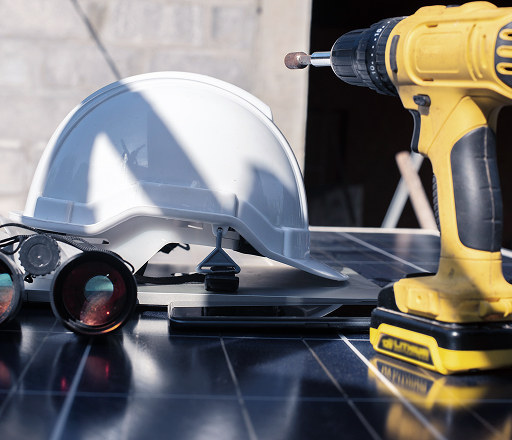 Close-up of a white safety helmet, yellow cordless drill, binoculars, and a tablet placed on a solar panel during installation work.