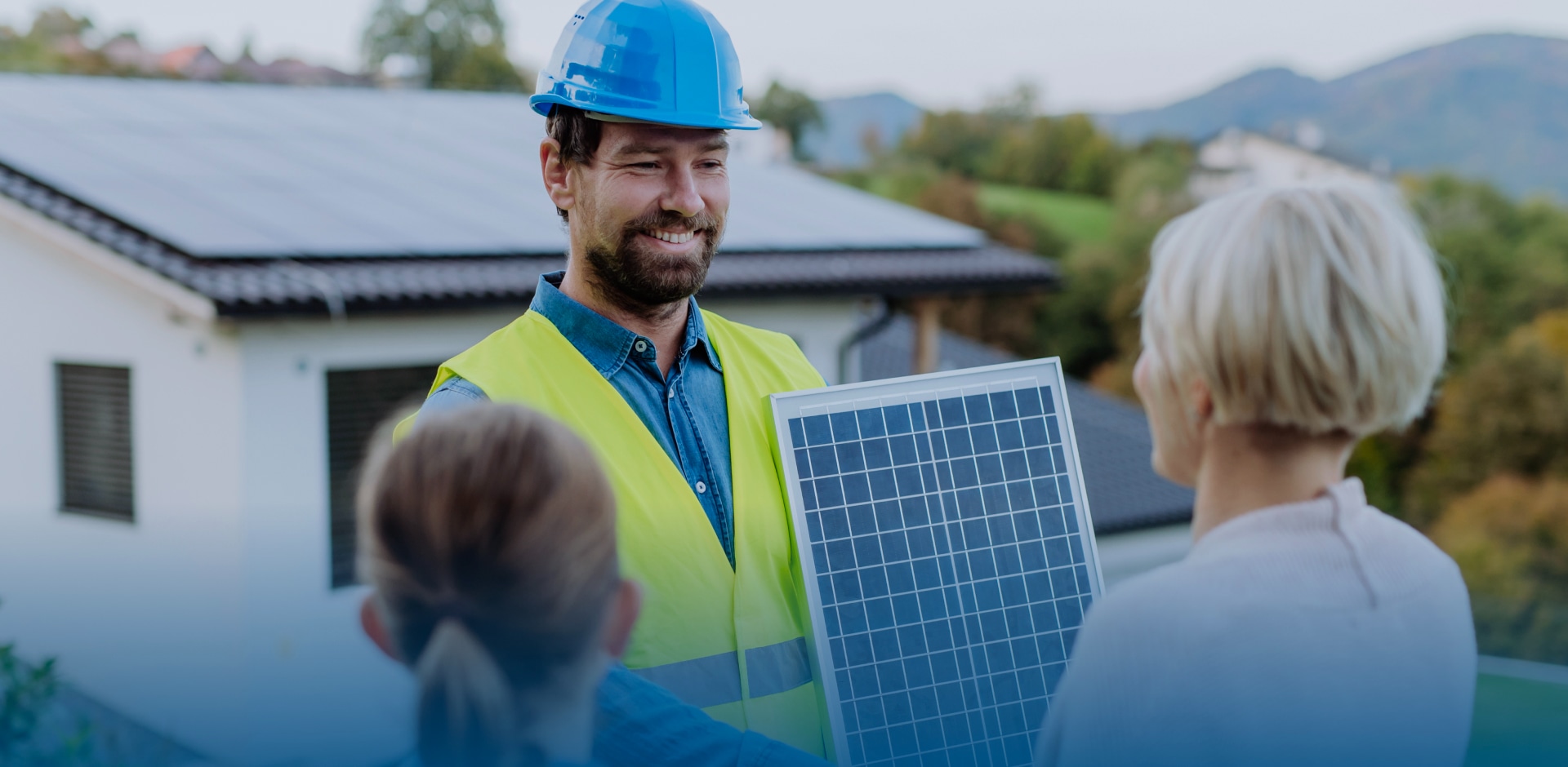 A smiling solar installer in a safety vest and blue helmet presenting a solar panel to two residential customers in front of a house with rooftop solar panels