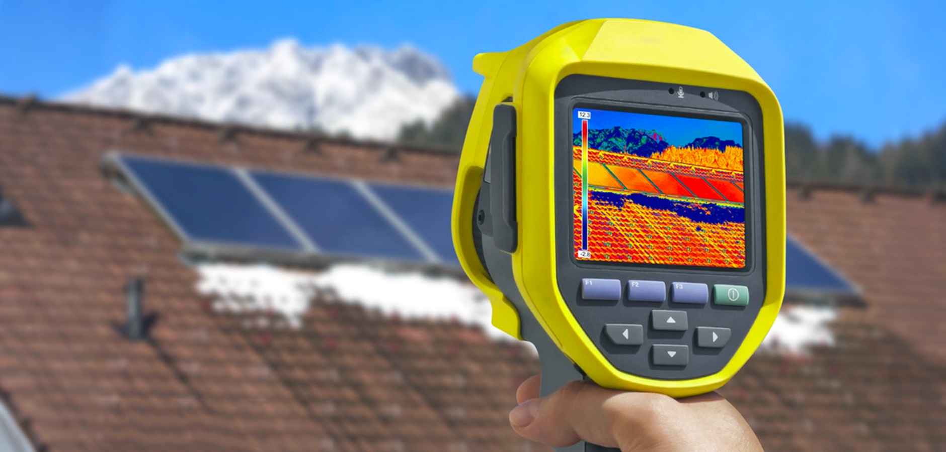 Handheld thermal imaging camera displaying a heat map of solar panels on a rooftop, showing temperature differences on the panels, with a clear view of solar modules in the background and a snowy mountain range